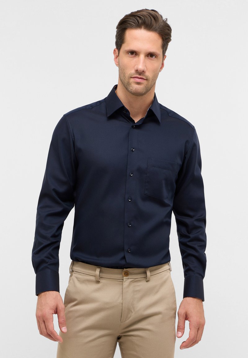 Navyblauw overhemd met lange mouwen en een button-down kraag, subtiele textuur, voorzak en zwarte knopen. G gedragen met lichtbeige broek.