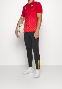 Homme portant un maillot de football AS Roma rouge et un pantalon noir, tenant un ballon de football blanc avec des motifs d'étoiles multicolores sur un fond blanc.