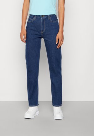 Osoba ubrana w ciemnoniebieskie jeansy o prostych nogawkach, jasnoniebieską bluzkę i białe trampki, stojąca na tle gładkiego, jasnego tła.