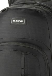 Zaino nero con tessuto strutturato, una tasca frontale con zip e un'etichetta con il logo "Dakine" ben in vista cucita sul materiale.