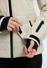 Lys beige pufferjakke med en blank finish, der har sorte accenter, lynlåse og velcro-manchetter. Tekstureret quiltet design på kroppen.