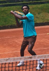 Light blue polo shirt, brown shorts, holding a tennis racket. White and black tennis shoes with a neon accent. Clay court background.