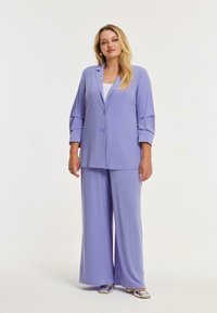 Vrouw in een lila wijde broekpak met een blazer met twee knopen en zilveren vierkante hakken, staand tegen een effen witte achtergrond.