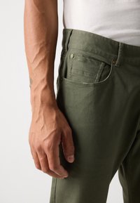 Des pantalons en coton verts présentent un design droit avec une texture lisse, des poches latérales et arrière, ainsi que des accents en métal.