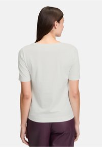 Femme aux cheveux bruns raides portant un haut blanc à manches courtes et un pantalon violet foncé, vue de dos sur un fond blanc.
