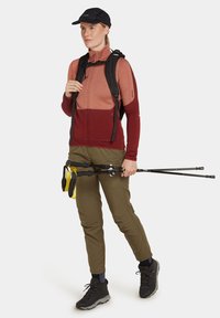 Camiseta de manga larga en tonos rojizos, pantalones ligeros en verde oliva, zapatos de senderismo negros, llevando bastones de trekking y equipado con mochila.