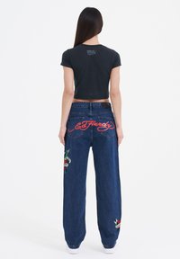 Black cropped t-shirt paired with blue denim pants featuring red embroidery and colorful graphic patches on the legs and back.
