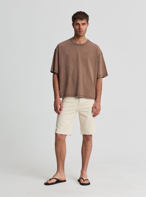 Giovane uomo con capelli corti e ricci, indossa una t-shirt marrone oversize, pantaloncini crema fino al ginocchio e infradito nere, in piedi su uno sfondo semplice.
