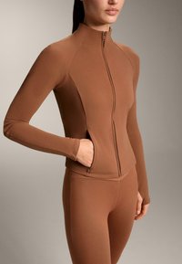 Giacca marrone aderente con zip realizzata in un materiale liscio ed elastico, caratterizzata da maniche lunghe, colletto alto e una tasca laterale.