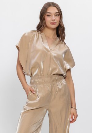 Vrouw draagt glanzende beige blouse met korte mouwen en bijpassende broek met elastische taille, poseert met één hand in de zak tegen een effen achtergrond.