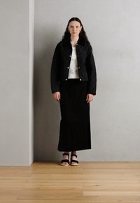 Veste matelassée noire avec des boutons-pression, portée sur un haut blanc et une longue jupe noire. Des sandales complètent la tenue. Fond uni et sol en bois.