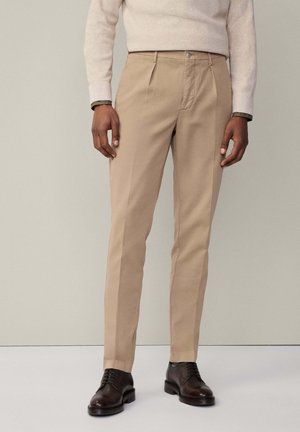 Beige tailored trousers with a straight leg design, featuring a button closure and side pockets, paired with brown dress shoes.