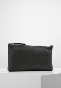 Pochette rectangulaire en cuir noir avec une fermeture éclair argentée sur le dessus, posée sur un socle blanc contre un fond gris clair uni.
