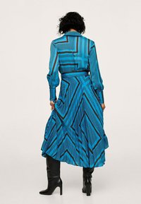 Vestido de manga larga en tela azul con rayas geométricas negras, que presenta una cintura ajustada, falda acampanada y un cuello. Llevado con botas negras hasta la rodilla.