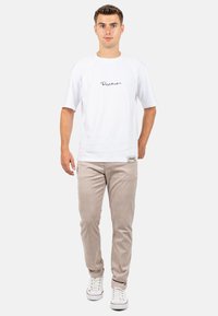 T-shirt blanc à manches courtes avec un logo noir sur la poitrine, associé à un pantalon en velours côtelé beige clair et des baskets blanches.