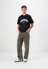 Svart bomull T-skjorte med "CORESPORT"-tekst, parret med løse, olivengrønne, falmede jeans og hvite sneakers med grå og svarte detaljer.