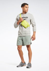Sudadera gris con el logo de Reebok, pantalones cortos verdes y un bolso bandolera amarillo brillante. El bolso presenta una textura suave y una correa ajustable.