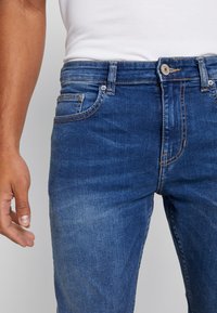 Modré džínové slim-fit kalhoty s vybledlým vzhledem, s klasickým pětipokovým designem, zapínáním na knoflíky a jemnými detaily švů.