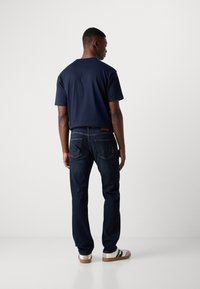 Tricou din bumbac bleu marin combinat cu blugi denim închisi. Detalii includ cusătura buzunarului din spate și adidași albi cu dungi negre.