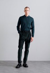Donkergroene polo met lange mouwen en ritskraag, gecombineerd met donkergroene op maat gemaakte broeken en zwarte stevige schoenen, op een neutrale achtergrond.