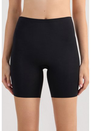 Frau trägt schlichte schwarze Kompressionsshorts bis zur Mitte des Oberschenkels und steht mit seitlich hängenden Armen vor neutralem Hintergrund.