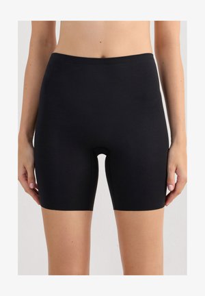 Kvinde iført almindelige sorte kompressionsshorts til midt på låret, stående med armene ned langs siderne mod en neutral baggrund.