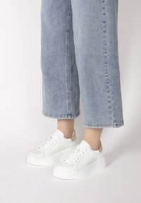 Weiße Sneaker mit einer strukturierten Oberfläche, abgerundetem Zehenbereich und kontrastierenden goldenen Akzenten an der Ferse, kombiniert mit hellblauen weiten Jeans.