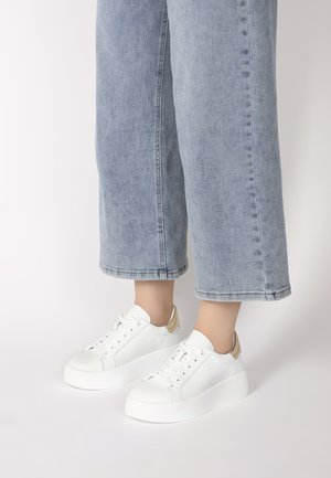 Weiße Sneaker mit einer strukturierten Oberfläche, abgerundetem Zehenbereich und kontrastierenden goldenen Akzenten an der Ferse, kombiniert mit hellblauen weiten Jeans.