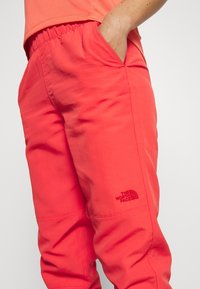 Korallinpunaiset housut, joissa on joustava vyötärönauha, sivutaskut ja pieni musta logo reidessä. Kangas näyttää kevyeltä ja sileältä.