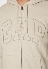 Sweat à capuche beige zippé avec un large logo "GAP" brodé dans une teinte légèrement plus foncée, avec une poche avant et une capuche à cordon.