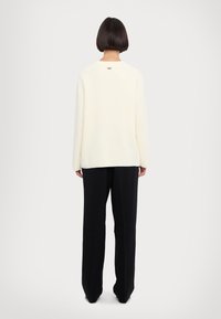 Pull en crème avec des côtes, à manches longues, col rond et un détail de logo au dos, associé à un pantalon noir ample.