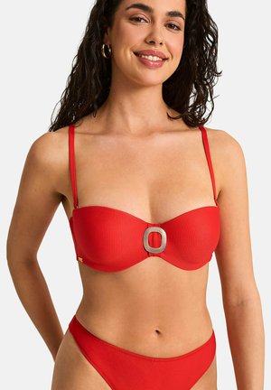 Femme portant un bikini rouge texturé avec une boucle rectangulaire dorée sur le haut, souriante avec des cheveux bouclés foncés et des boucles d’oreilles anneaux dorés.