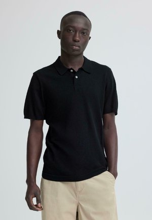 Giovane uomo in piedi davanti a uno sfondo semplice, indossa una polo nera a maniche corte e pantaloni beige con una mano in tasca.