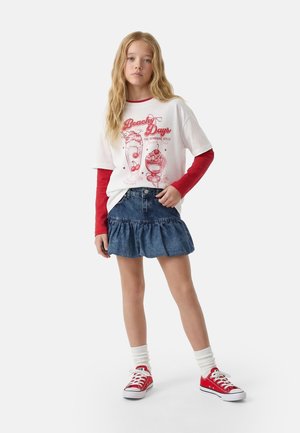 Chica rubia con camiseta blanca con el gráfico "Beach Days" sobre mangas largas rojas, falda azul de mezclilla con volantes, calcetines blancos y zapatillas deportivas rojas.