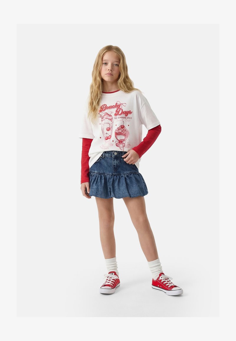Fille blonde portant un t-shirt blanc imprimé « Beach Days » par-dessus des manches longues rouges, une jupe en jean bleue à volants, des chaussettes blanches et des baskets rouges.