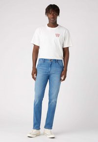Joven con dreadlocks cortos que lleva una camiseta gráfica blanca, jeans de un azul claro y zapatillas altas blancas, de pie sobre un fondo sencillo.