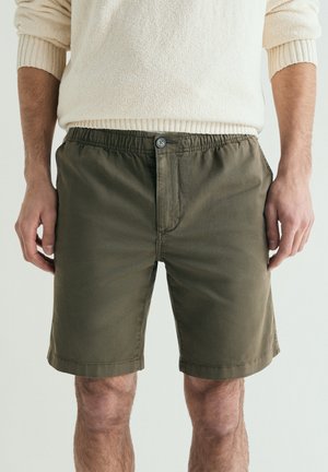 Hombre con pantalones cortos verdes oliva con botones y un suéter crema con las mangas remangadas, de pie frente a un fondo liso.