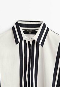 Blouse rayée avec des rayures verticales noires et blanches, comportant un col pointu et une patte de boutonnage avec des détails de boutons. Tissu léger.