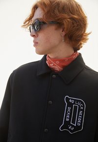 Black fleece jacket with button closure, featuring a white embroidered patch with "NO LUCK NEEDED DICKIES" and a textured bandana scarf underneath.