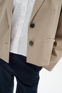 Beiger Blazer mit schwarzen Knöpfen über einem weißen Hemd, kombiniert mit dunkelblauen Hosen.