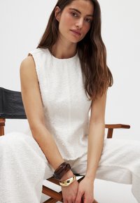 White textured sleeveless top with a round neckline, paired with matching pants. Accessories include a dark and a gold bangle bracelet.