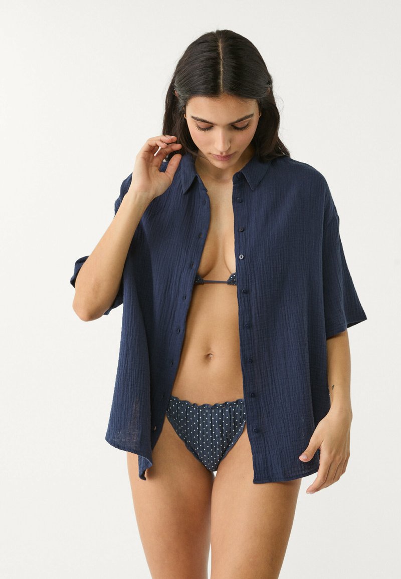 Camicia blu navy a maniche corte in tessuto testurizzato, di taglio rilassato; indossata sopra un bikini a pois bianchi.