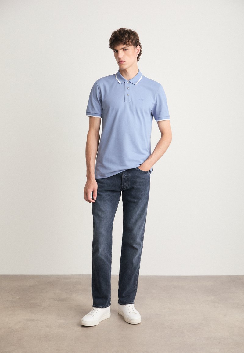 Polo shirt bleu clair avec col et bord blanc, patte de boutonnage à trois boutons, associé à un jean foncé et des baskets blanches. Texture unie, coupe décontractée.