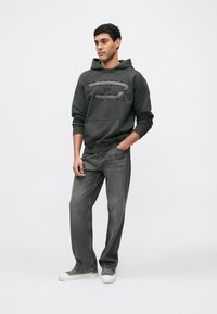 Temno siva jopa s kapuco in sprednjim žepom ter grafičnim napisom na prsih, kombinirana z rahlo širokimi sivimi kavbojkami in supergami z zelenimi poudarki.