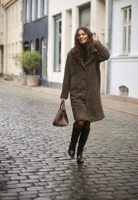 Palton mare, pufos, de culoare maro, cu închidere pe butoane, purtat cu cizme negre până la genunchi. Ținând o mică geantă maro. Fundal cu stradă de piatră cubică.