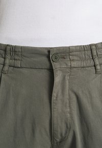 Pantalons kaki avec une texture lisse, dotés d'une fermeture à bouton et d'une taille élastique pour un ajustement parfait. Détails cousus sur les côtés.