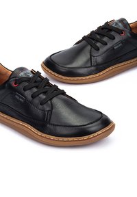 Zapatillas negras de cuero con textura suave, que cuentan con cordones negros, suelas de goma en contraste color beige y sutiles acentos rojos en la marca.
