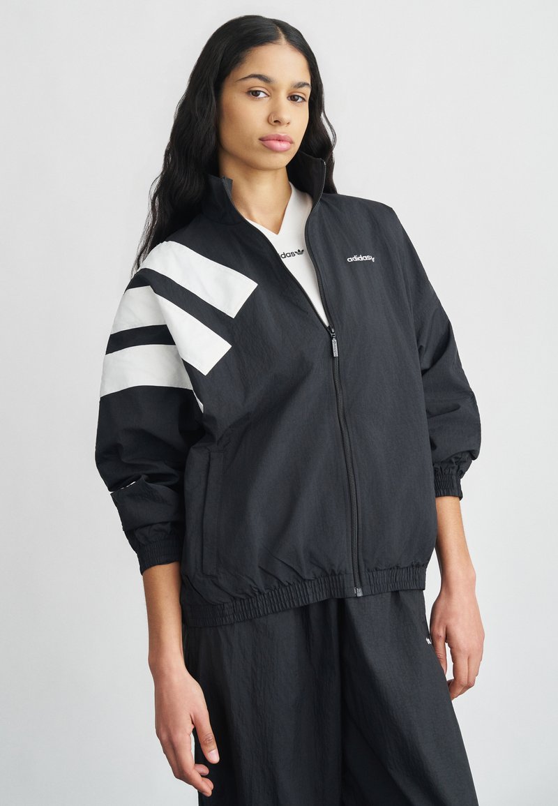 Sort glidelåsjakke med hvite aksentstriper og logo. Har elastiske mansjetter og linning, laget av lett materiale.