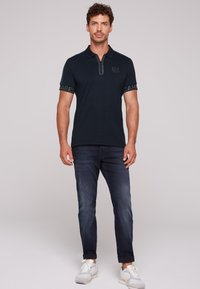 Granatowa koszulka polo z zamkiem przy kołnierzyku, z szarym logo na piersi i akcentami "CAMEL" na rękawach, w połączeniu z ciemnymi dżinsami i trampkami.