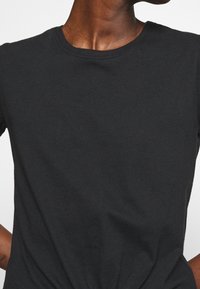 Øverste del af kroppen på en person iført en ensfarvet sort crewneck t-shirt, med den ene arm let bøjet og synlig tatovering på håndleddet.